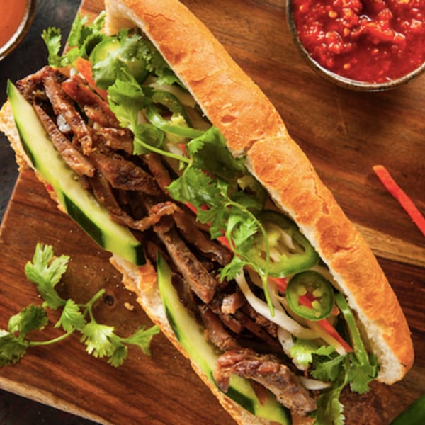 A Bon Mi Vietnamese sandwich with meat, cucumbers, cilantro, and jalapeños on a wooden board.